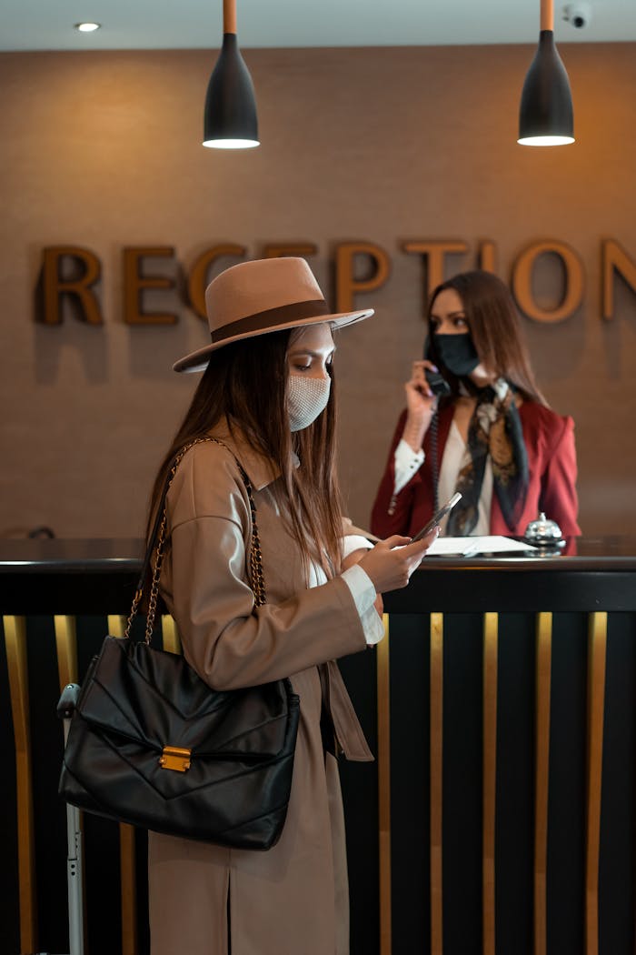 Woman in face mask at hotel reception counter checking her phone, symbolizing the new normal.